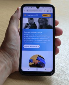 A mobile phone held in the palm of a hand showing Recovery College Online website on the screen