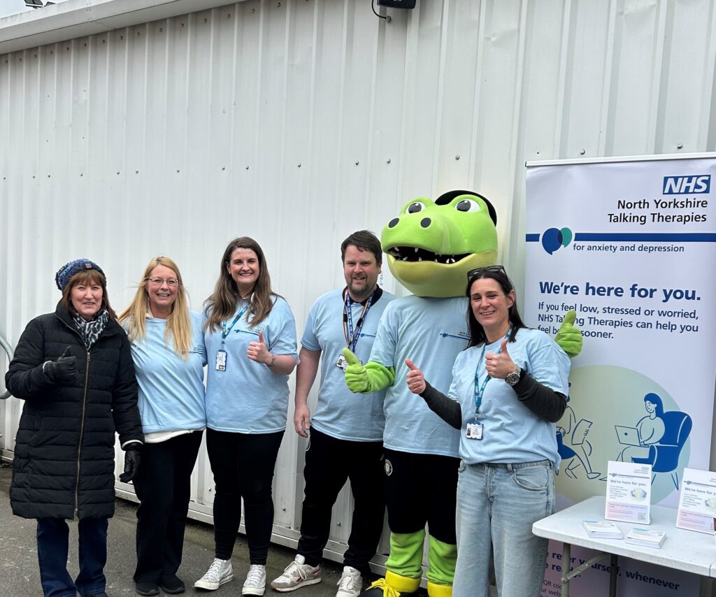 North Yorkshire Talking Therapies staff members and Harrogate Town AFC's mascot smiling at the camera next to the service banner at the gates of the stadium.