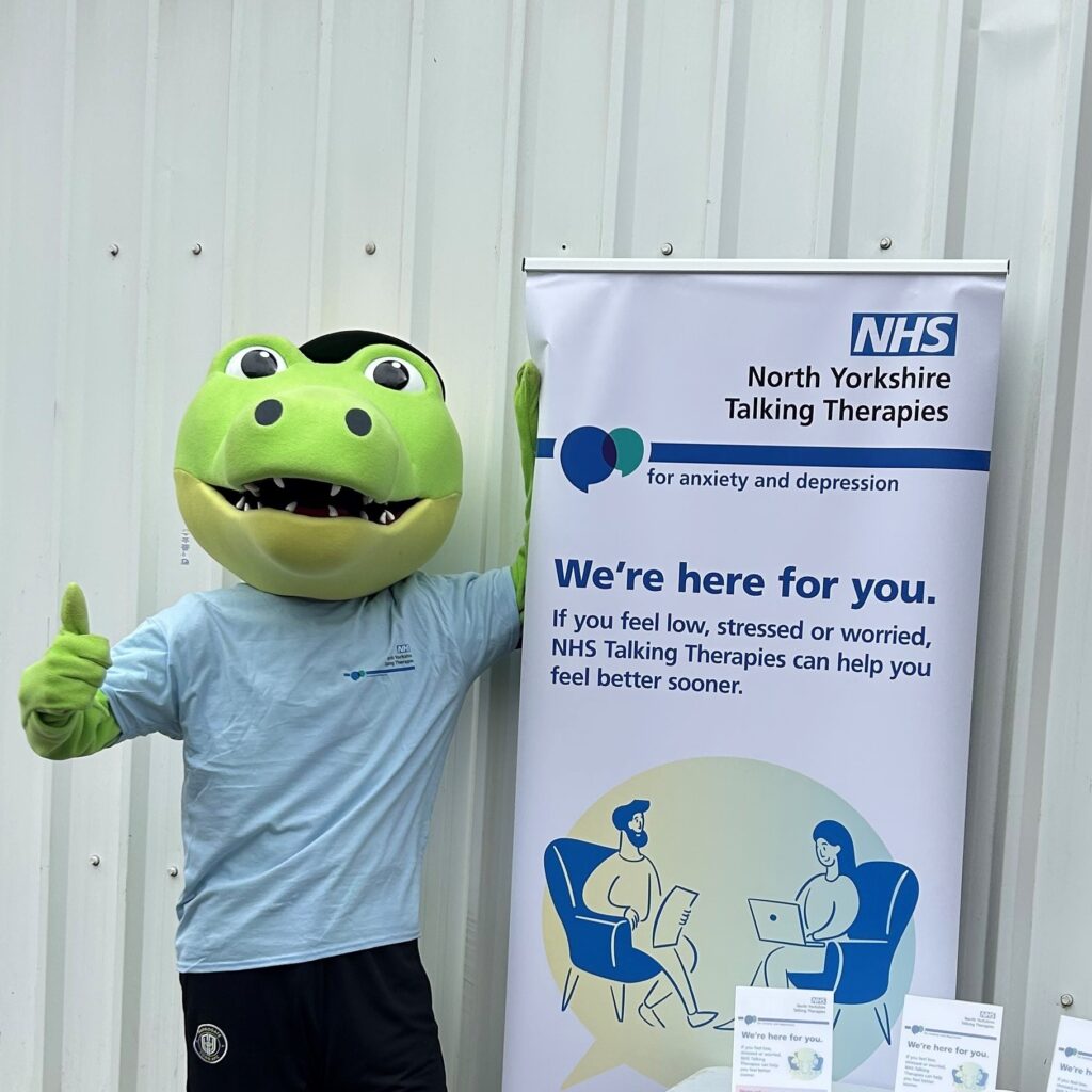 Harrogate Town AFC's masco, Harry Gator, stood next to North Yorkshire Talking Therapies' banner.
