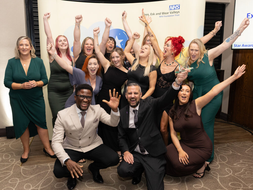 Durham and Darlington perinatal mental health service gathered in front of a yellow Star Awards banner. They are cheering with their arms in the air.