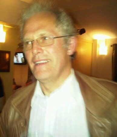 Man with grey hair and glasses wearing a white shirt and brown jacket