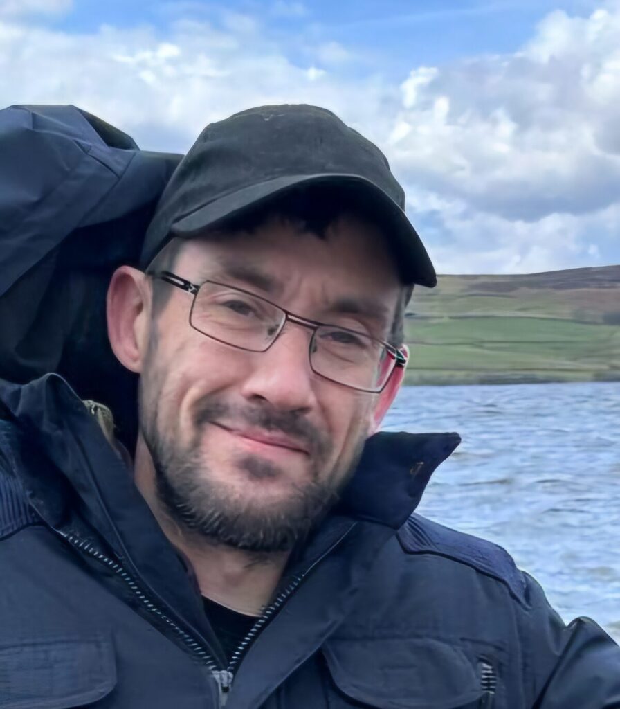 Man with glasses and a beard wearing a cap, coat and stood in front of a lake