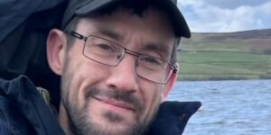 Man with glasses and a beard wearing a cap, coat and stood in front of a lake